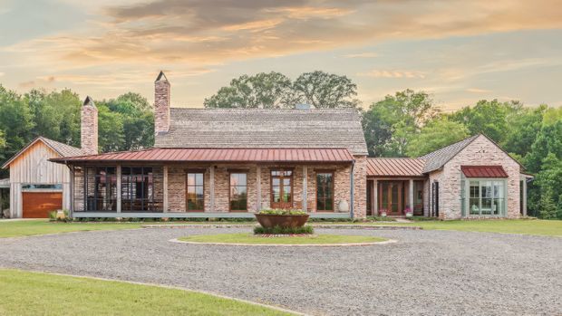 A wide exterior view of a low-rise country-style residence arranged around a broad gravel forecourt. The main brick house has a long pitched roof, multiple brick chimneys, a covered glazed veranda, timber-framed windows and doors, and a red-brown metal roof over the veranda. Smaller attached or nearby wings include a gabled timber-clad structure and a brick garden-room-like volume. Mature trees and lawns surround the property.