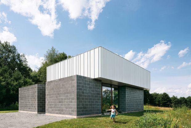 A compact modern, single-storey building set in a grassy, tree-lined rural or semi-rural landscape. The lower walls are constructed from dark grey concrete masonry blocks, while the upper volume is clad in vertical white metal panels with a simple pitched or folded roof form. Large floor-to-ceiling glazed openings sit beneath the projecting upper volume, creating a strong geometric composition. A child stands near the entrance, providing scale.
