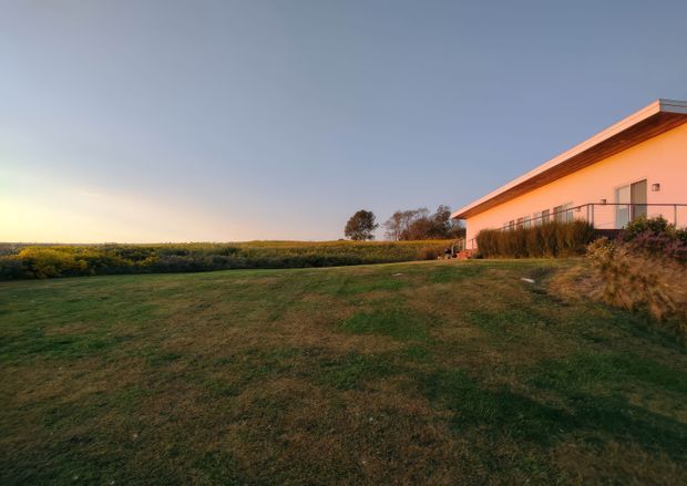 A low, modern residential building viewed from a broad grassy lawn at sunset. The simple rendered facade has a shallow projecting roof with dark trim, a long balcony or raised walkway with metal railing, and large glazed openings. Dense hedges and open rural fields extend across the background, creating a quiet, expansive setting.