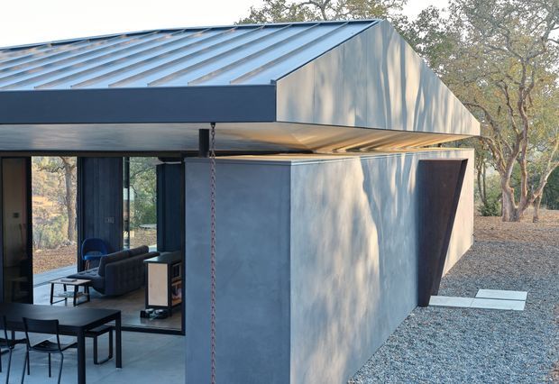 Exterior view of a low-slung contemporary building in a wooded, gravel-covered setting. The structure has a dark standing-seam metal gable roof, angular folded roof and wall forms, smooth grey-blue cladding, large dark-framed glazed openings, and a covered outdoor dining area. A living room with dark seating is visible through the glazing.