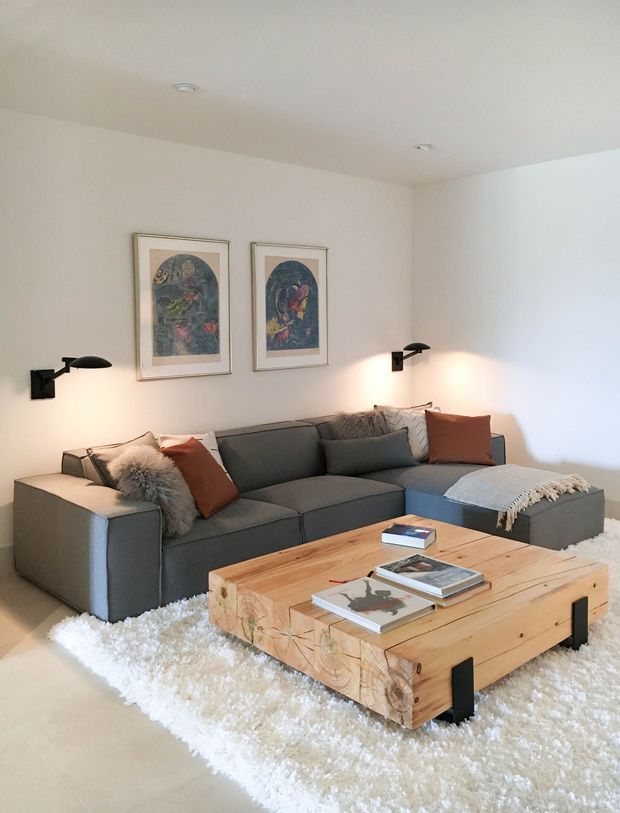 A bright, minimalist living room with a large charcoal-grey sectional sofa, rust and neutral cushions, and a substantial rustic timber coffee table with black metal legs. A white shag rug softens the pale floor, while two framed artworks and sleek black wall sconces decorate the off-white wall.