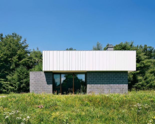 Front exterior view of a compact modern single-storey building set within a natural meadow and surrounded by mature trees. The structure has a simple rectangular form, a white vertically panelled upper volume projecting over dark grey concrete masonry walls, and a central glazed entrance or window wall with dark frames. A low-profile roof element is visible above the upper volume.