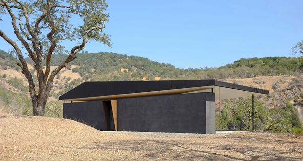 A low, minimalist dark-clad building set within a dry, hilly landscape. The structure has a simple flat or very low-pitched roof with deep overhangs, dark textured walls, and a narrow timber-toned entry or recessed opening. Mature trees and rugged hills surround the building under a clear blue sky.