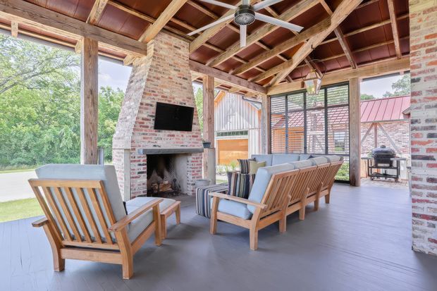 A spacious covered outdoor living pavilion with an exposed timber roof structure, brick fireplace and chimney, ceiling fan, timber-framed glazing, and a comfortable seating arrangement facing the fireplace and television. The room opens to a green garden and includes a barbecue area visible at the rear. The design combines rustic structural materials with a relaxed contemporary outdoor entertaining setup.
