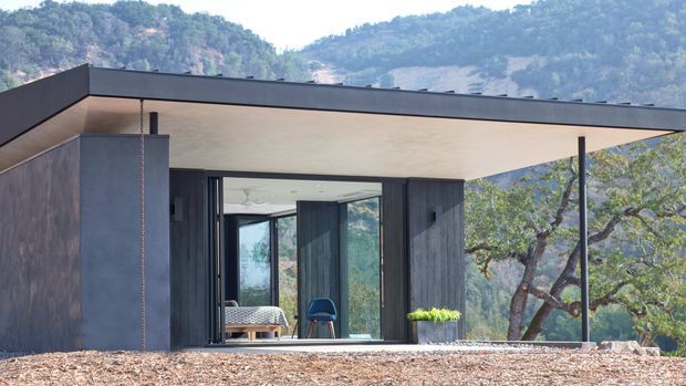 A modern single-storey pavilion-like building set against a wooded mountainous landscape. The structure has a broad flat roof with deep cantilevered eaves, dark vertical cladding, extensive floor-to-ceiling glazing, and a sheltered outdoor terrace. A bedroom with a bed and chair is visible through the open glazing.