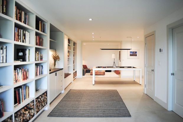 Bright, contemporary open-plan interior viewed from a hallway or entry area. Floor-to-ceiling white built-in bookshelves and display joinery line the left wall, leading toward a softly furnished living area with a low white table, sofa, artwork, and floor lamp. A large neutral striped rug defines the foreground, while white paneled doors and recessed ceiling lighting reinforce the clean, refined aesthetic.