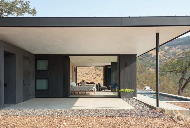 A modern single-storey hillside residence viewed from an exterior covered patio or terrace. The composition features a broad flat roof with a slim dark metal fascia, dark vertical cladding, expansive sliding glass openings, and a minimalist concrete terrace edged with gravel. A living room is visible through the open glazing, with a swimming pool and rugged landscaped hillside beyond.
