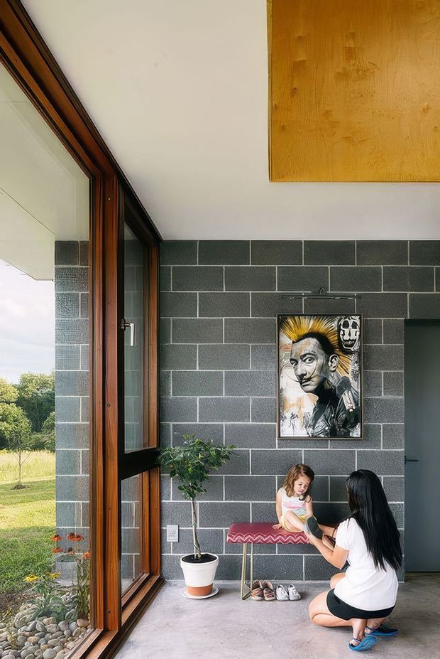 A bright modern entryway with floor-to-ceiling timber-framed glazing on the left, a dark grey block feature wall, polished concrete floor, and a simple timber ceiling panel. A small bench, potted tree, artwork, and family members occupy the space. The glazing opens toward a landscaped garden.