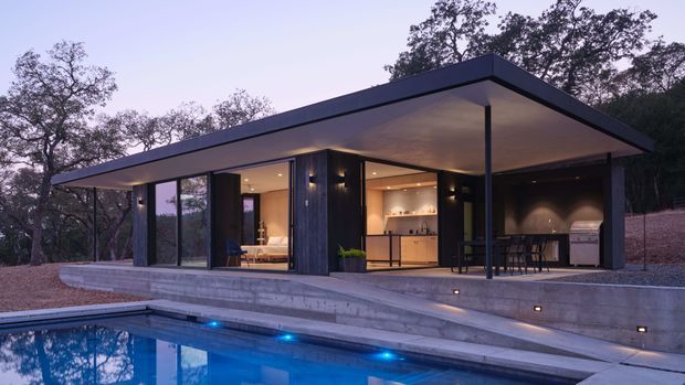 A contemporary flat-roof pavilion overlooking a swimming pool in a wooded setting. The open-sided structure combines dark vertical cladding, extensive floor-to-ceiling glazing, warm recessed lighting, a compact kitchen or bar area, outdoor dining, and a covered barbecue zone. Broad concrete terraces step down to the illuminated pool.