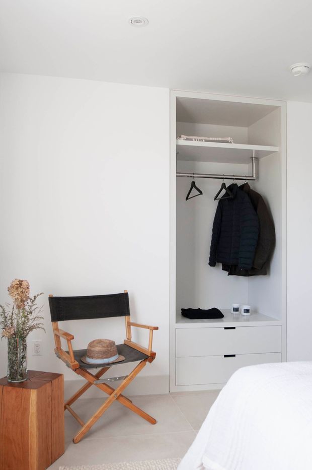 Bright, minimalist bedroom corner with a built-in open wardrobe and drawers, showing hanging clothes, a director-style wooden chair, a small wooden side table with dried flowers, and part of a neatly made bed. The room has white walls and ceiling, pale tiled flooring, and understated contemporary detailing.