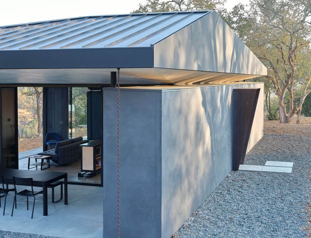 Exterior view of a contemporary single-storey building with a sharply folded pitched metal roof and projecting geometric facade volumes. Dark metal cladding and framing contrast with pale concrete or rendered wall surfaces. Large glazed openings reveal a modern living and dining area, while the surrounding gravel landscape and mature trees create a secluded natural setting.