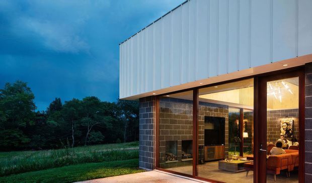 Twilight exterior view of a contemporary two-storey building set beside a wooded landscape. The lower level uses dark masonry block walls with extensive floor-to-ceiling glazing, warm timber framing, and a sheltered living area visible inside. The upper level is clad in pale vertical metal panels with a projecting cantilevered form. Warm interior lighting contrasts with the cool blue evening sky.