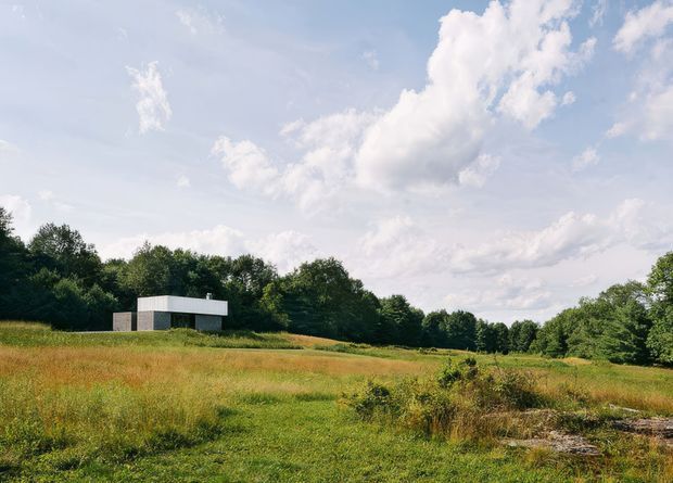 A minimalist, single-storey flat-roof building set within an expansive meadow, viewed from a distance. The structure has crisp white horizontal roof and wall elements with dark glazing or openings, surrounded by trees and tall grasses under a bright partly cloudy sky.