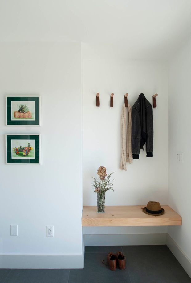 A bright, minimalist entryway with white walls, a built-in light timber bench, and four wall-mounted coat hooks holding outerwear and a scarf. Framed artwork hangs on the adjacent wall, while shoes sit beneath the bench and a vase of dried flowers and a hat rest on top. The space has a dark grey tiled floor and a clean, functional composition.