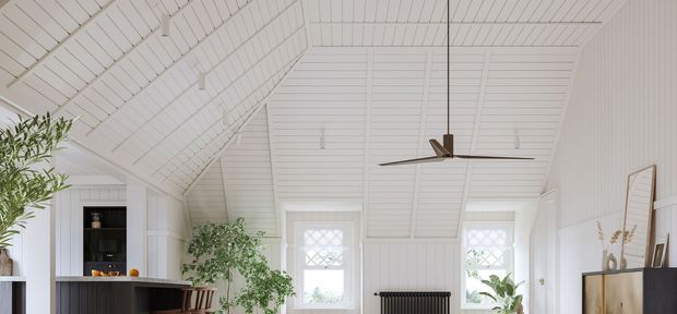 Bright white interior with a steep vaulted, gabled ceiling clad in narrow horizontal boards and matching wall panelling. Decorative grilles appear on the tall windows, while a long dark ceiling fan hangs centrally. The lower portion shows partial dark cabinetry and shelving, indoor plants, a radiator, and a sideboard, suggesting an airy living or kitchen-adjacent space.