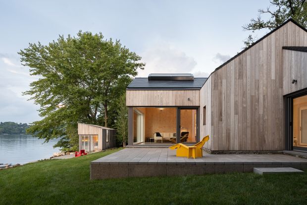 Exterior view of a contemporary lakeside home with a low, angular gable-roofed form clad in vertical pale timber boards. Large glazed openings connect the interior to a broad paved terrace furnished with a bright yellow chair. A smaller matching outbuilding sits near the water, with mature trees and the lake forming the surrounding setting.