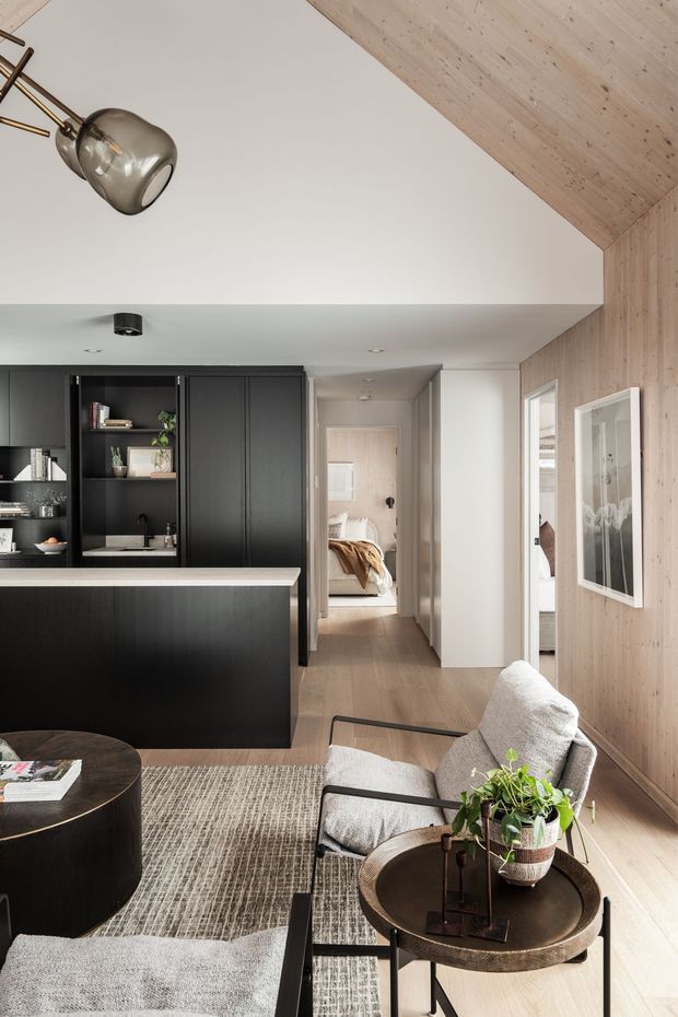 A refined open-plan apartment interior combining a black kitchen with integrated shelving and island, a compact living area, and a hallway leading to a bedroom. Light timber wall and ceiling cladding, pale timber flooring, white walls, dark joinery, and contemporary furnishings create a warm, sophisticated look. A sculptural pendant light, framed artwork, textured rug, upholstered armchairs, and round coffee tables complete the space.