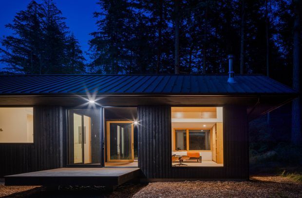 A compact single-storey cabin-like house photographed at dusk in a wooded setting. The dark vertically clad facade has a low-pitched metal roof, large warm-lit windows, a glazed entry door, and a small projecting timber deck. Interior furnishings are visible through the glazing, including an orange sofa or chair and a desk-like surface.