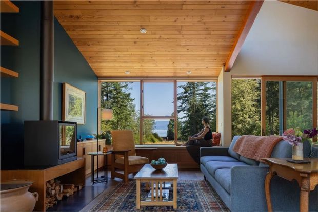 A warm, timber-lined living room with a vaulted ceiling and exposed beam, large timber-framed windows, and a scenic water view beyond the trees. The room includes a blue sofa, patterned rug, wood-burning fireplace or stove with built-in firewood storage, lounge chair, coffee table, and dark feature wall shelving. The interior has a relaxed mid-century and rustic character with abundant natural light.
