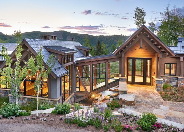 Exterior view of a multi-wing mountain home with steep gabled roofs, dark timber framing, warm wood siding, stone masonry bases, and extensive glazed connectors. The landscaped foreground includes planted beds and natural stone paths, while forested hills rise behind the property at dusk.