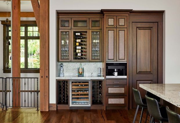 A richly detailed traditional-style home bar and drinks joinery wall, featuring dark timber cabinetry with glazed display doors, wine storage, a wine refrigerator, glassware shelving, and decorative leaded-glass panels. The built-in joinery sits beside a paneled timber door and opens toward a dining or kitchen area with a stone benchtop and stools. Exposed timber posts, a timber balustrade, and polished timber flooring add warmth and architectural character.