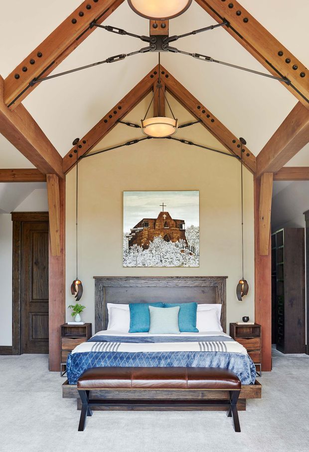 A symmetrical bedroom with a prominent timber post-and-beam vaulted ceiling, exposed trusses, dark metal tension rods, and two warm pendant lights. The room features a centered bed with a weathered timber headboard, blue and white bedding, matching bedside tables, a leather bench, dark timber doors and wardrobes, a grey carpeted floor, and a rustic architectural painting above the bed.