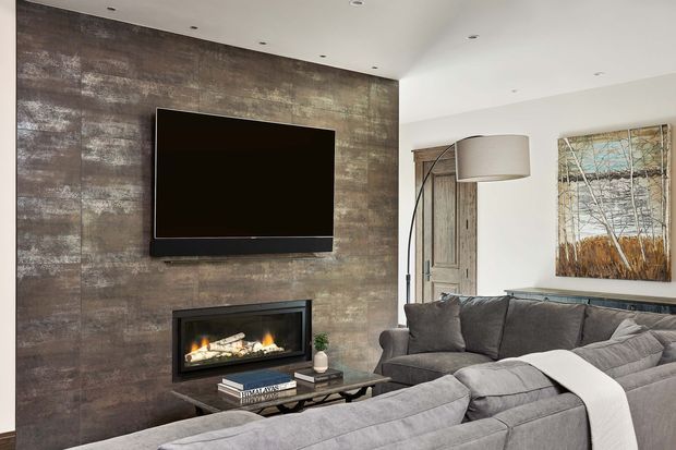 Contemporary living room featuring a large grey sectional sofa, wall-mounted television, linear fireplace, dark textured feature wall, coffee table, floor lamp, and a rustic timber door. The room has recessed ceiling lighting and a neutral grey, white, and brown palette.