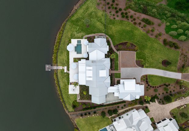 Aerial top-down view of a large waterfront residential property with several connected buildings featuring light-coloured pitched and gabled roofs. The landscaped site includes curved driveways, planted garden beds, lawns, and a private dock extending into the water.