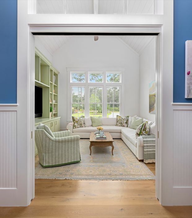 A bright, refined sitting room viewed through a wide decorative doorway. The room has a white vaulted or pitched beadboard ceiling, large multi-pane windows with white plantation shutters, pale green built-in cabinetry and shelving, and a light upholstered seating arrangement around a traditional wooden coffee table. A large muted patterned rug sits over wide-plank timber flooring. White wall panelling and trim frame the opening, with deep blue wall sections visible in the foreground.