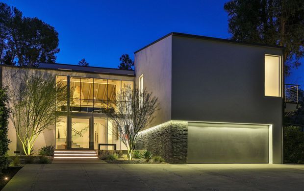 Twilight exterior view of a contemporary two-storey residence with a box-like flat-roof form, smooth light rendered walls, extensive glazed entry facade, illuminated stair hall, landscaped frontage, and an integrated double garage. A textured dark stone-clad wall anchors the garage level, while warm interior and exterior lighting contrasts with the deep blue sky.