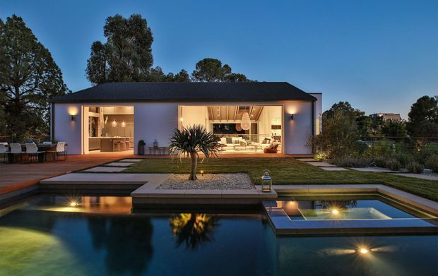 A contemporary single-storey residence photographed at dusk, with a dark low-pitched roof, pale rendered exterior walls, and expansive illuminated openings revealing an open-plan interior. A landscaped lawn and paved paths lead to a geometric reflecting or swimming pool in the foreground, with integrated lighting, a raised shallow section, and a central sculptural planting bed. Mature trees frame the property.
