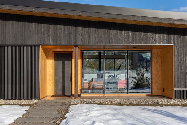 Front exterior view of a low-slung contemporary building in a snowy setting. The facade combines dark vertical timber cladding, a broad flat roof with exposed timber eaves, black-framed glazed entry doors and windows, and a warm timber-lined recessed entrance. A gravel strip and stepping-stone path lead through snow to the entry.