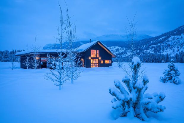 A warmly lit timber-clad mountain cabin set in a snow-covered landscape at blue hour. The low pitched roof, dark exterior, glowing windows, young deciduous trees, snow-laden evergreen shrubs, and forested mountainous backdrop create a secluded alpine setting.