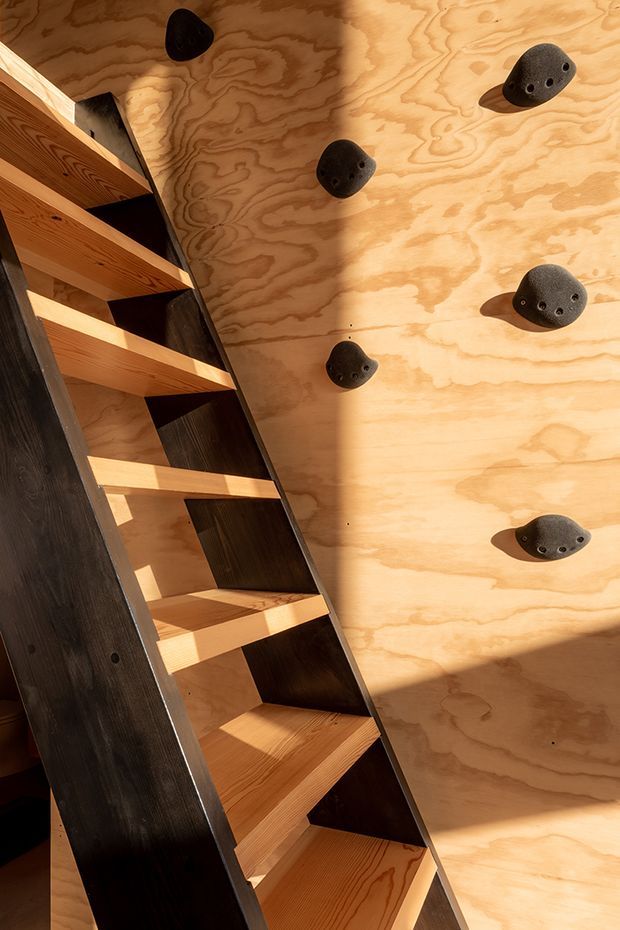 Interior view of a compact climbing wall beside a steep timber stair or ladder structure. The wall is clad in warm plywood with dark climbing holds, while the stairs use exposed timber treads and dark side panels. Strong sunlight creates pronounced shadows across the wood surfaces.