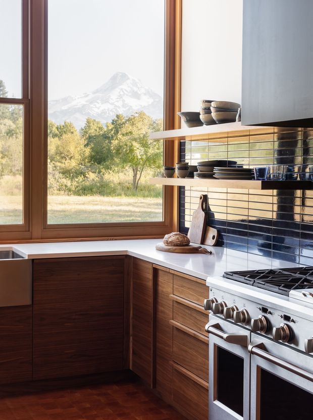 A warm, refined kitchen with dark timber cabinetry, white countertops, open timber shelves, and a glossy dark blue tiled splashback. A large stainless-steel range cooker sits beneath a concealed hood. Full-height timber-framed windows provide views of trees and a snow-capped mountain, while ceramic servingware and chopping boards add restrained styling.