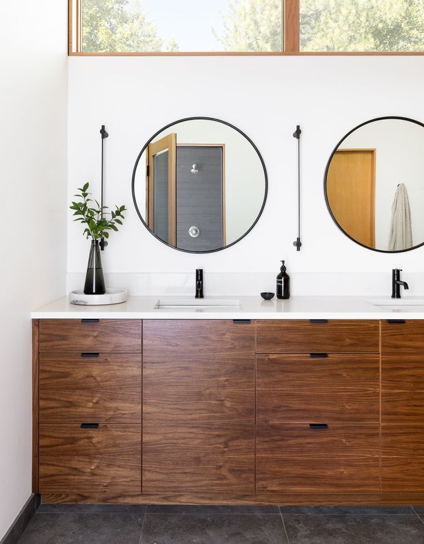 A bright, contemporary bathroom vanity with a long wall-mounted timber cabinet featuring rich dark walnut-toned grain and minimalist black pulls. A white benchtop contains two integrated basins with slim black tapware, complemented by two large round black-framed mirrors. The white wall is accented by slim black vertical wall lights, while a high timber-framed window provides natural light. Dark grey floor tiles and a small potted plant complete the clean, refined composition.