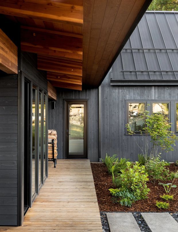 A covered timber deck and entry walkway within a modern dark-clad residential courtyard. The building features charcoal vertical cladding, extensive dark-framed glazing, warm exposed timber soffits and decking, and a planted garden bed with stepping slabs and dark gravel. The composition emphasizes the sheltered outdoor circulation space and contrasting natural materials.