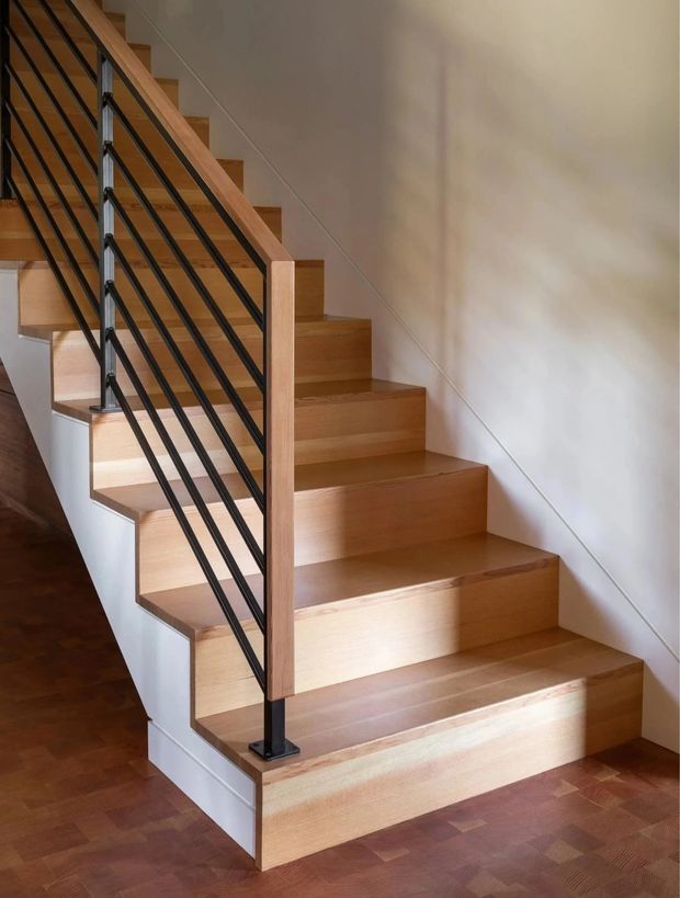 Interior photograph of a straight staircase with warm timber treads and risers, white walls, and a black metal balustrade with slim horizontal rails and timber posts. The staircase rises from a timber-floored lower level and has a clean, contemporary minimalist appearance.
