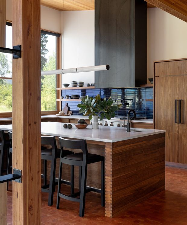 A warm, refined kitchen with a large central island and seating for three. The space features rich timber structural posts, ceiling lining, cabinetry and island cladding, a pale stone island benchtop, dark glossy blue horizontal tile splashback, a sculptural dark rangehood, black-framed glazing, and a patterned timber parquet floor. Black bar stools and a large leafy plant add contrast. The kitchen opens toward a bright dining or living area visible at the left.