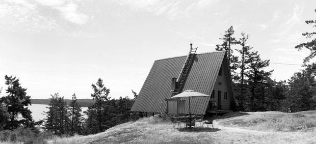 Black-and-white exterior photograph of a steeply pitched A-frame cabin with ribbed metal roofing, timber-clad walls, and a small elevated deck or balcony. The cabin sits among trees on a sloping, rugged site with a distant body of water visible beyond.
