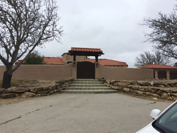 Exterior view of a gated, low-rise property with stucco boundary walls, terracotta tiled roofs, a dark timber entrance gate, patterned tiled steps, and rustic stacked-stone landscaping walls. Leafless trees and an overcast sky surround the entrance.
