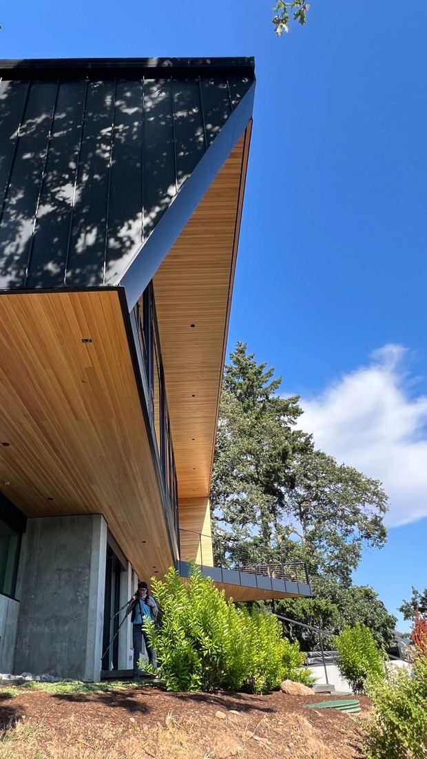 Upward-looking exterior view of a contemporary multi-level house with a dramatic projecting roof, dark standing-seam metal roofing, warm timber soffits, concrete walls, extensive glazing, and a cantilevered balcony with dark metal railing. The home is set among landscaped planting on a sloping site beneath a clear blue sky.