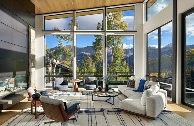 A bright, contemporary living room with expansive floor-to-ceiling glazing overlooking a dramatic mountainous landscape. The room features a high timber-lined ceiling, dark built-in cabinetry, a pale sectional sofa, sculptural lounge chairs, a round coffee table, and a large abstract-patterned rug. Natural light, warm timber, and panoramic views create an airy yet refined atmosphere.