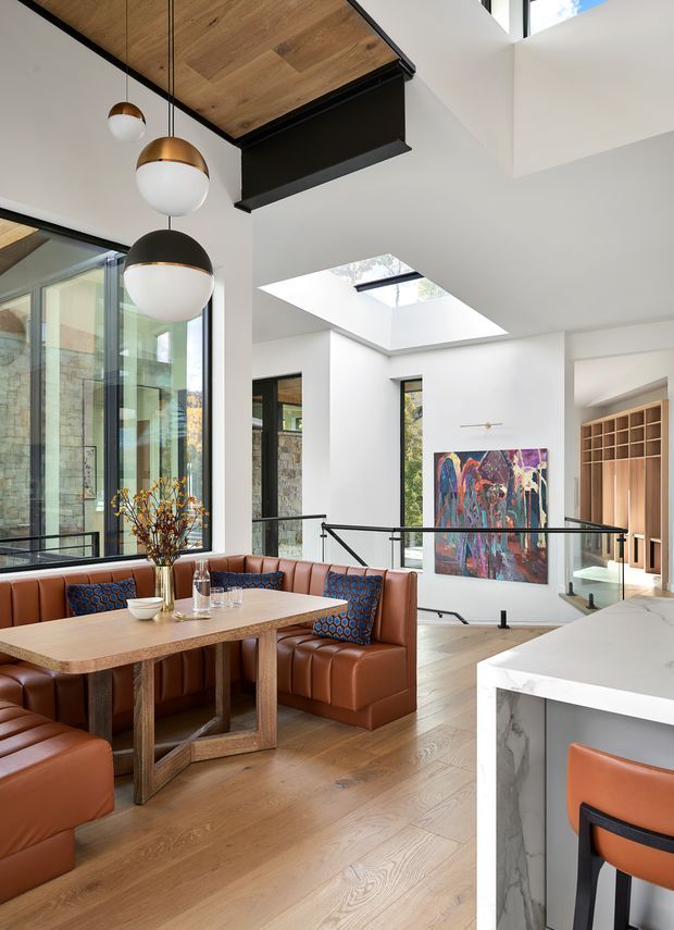 A bright, contemporary open-plan kitchen and dining area overlooking a double-height stairwell and lower-level space. The room features a built-in tan leather banquette, matching upholstered dining chairs, a large timber dining table, light timber flooring, white walls, expansive glazing, a skylight, black metal balustrades, spherical pendant lights, and a white stone kitchen island with waterfall edging. A colourful abstract artwork and timber wine or display joinery are visible beyond the dining area.