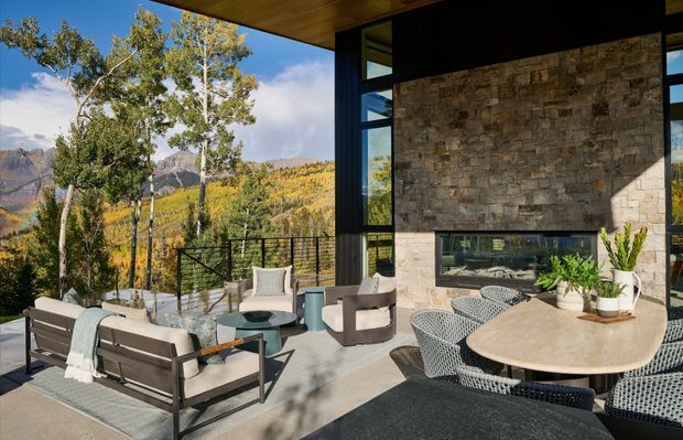 A contemporary mountain-view outdoor terrace adjoining a glass-walled residence. The covered patio features a dining table with woven chairs, a lounge seating arrangement, concrete paving, a dark metal balustrade, and a prominent natural stone-clad wall with a horizontal window. Aspen trees and rugged mountains are visible beyond the terrace.