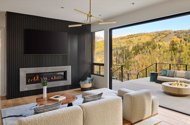 A refined contemporary living room opening onto a spacious terrace, with expansive black-framed glazing overlooking a wooded mountain landscape. The room features a dark vertical slatted feature wall, wall-mounted television, linear gas fireplace with a pale stone surround, light timber flooring, sculptural gold ceiling light, and textured cream seating arranged around a low timber coffee table. Outdoor lounge seating and a fire table are visible on the terrace.