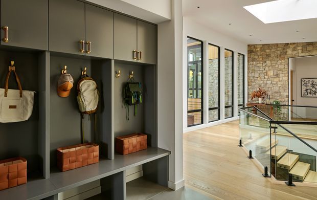 A refined residential entryway with dark built-in mudroom-style cabinetry, open cubbies, brass hardware, hooks, bags and woven storage baskets. The entry opens onto a bright circulation area with pale timber flooring, full-height black-framed glazing, a skylight, a glass balustrade and a staircase. A textured stone feature wall is visible in the background.