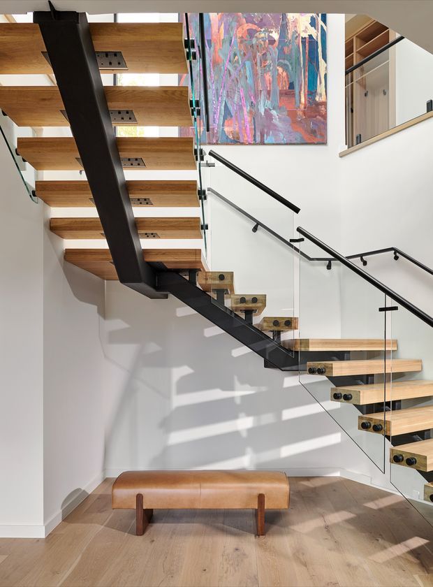 A contemporary interior stairwell featuring an open-riser, multi-flight staircase with thick natural timber treads, a dark painted steel structural spine and slender black handrails. Clear glass balustrades preserve the open feel. White walls, pale timber flooring, built-in timber shelving and a colourful abstract artwork create a bright, gallery-like setting. A low brown leather bench sits beneath the stairs.