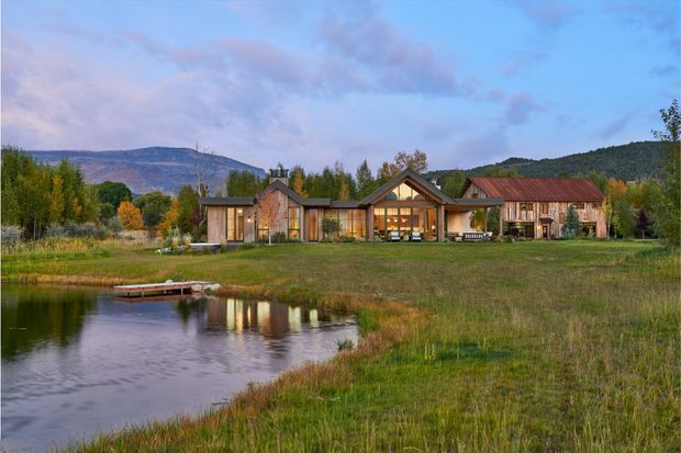 Exterior view of a low-rise rural residence beside a pond, set in a broad grassy landscape with wooded hills and autumn vegetation beyond. The home combines dark timber or metal-clad gabled volumes, warm timber-framed glazing, and a prominent central glazed gable, with a long rust-toned roofed wing to the right. A small dock projects into the pond in the foreground.