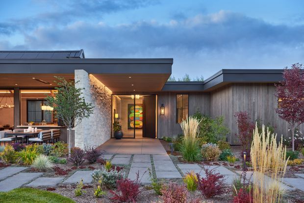 Single-storey contemporary residence with a low-profile flat roof, dark metal roof edging, extensive glazing, and a mix of vertical timber cladding and pale textured masonry. The front entry opens onto a paved path through a landscaped, drought-tolerant garden. A covered outdoor dining or living area is visible to the left, with warm interior lighting and modern built-in elements.